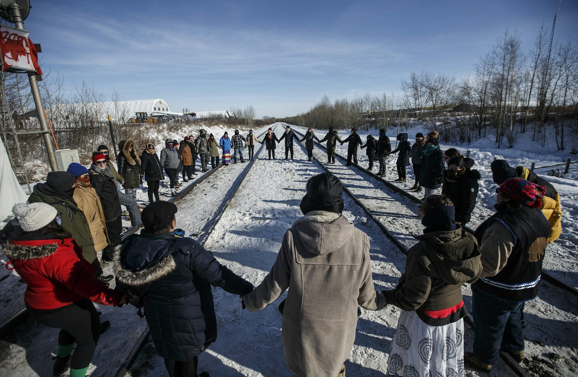 A group of people hold hands in a circle around a train track. 