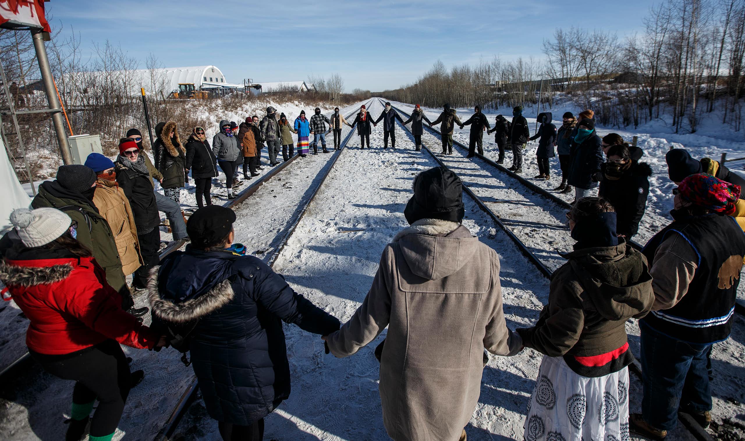 A group of people hold hands in a circle around a train track. 