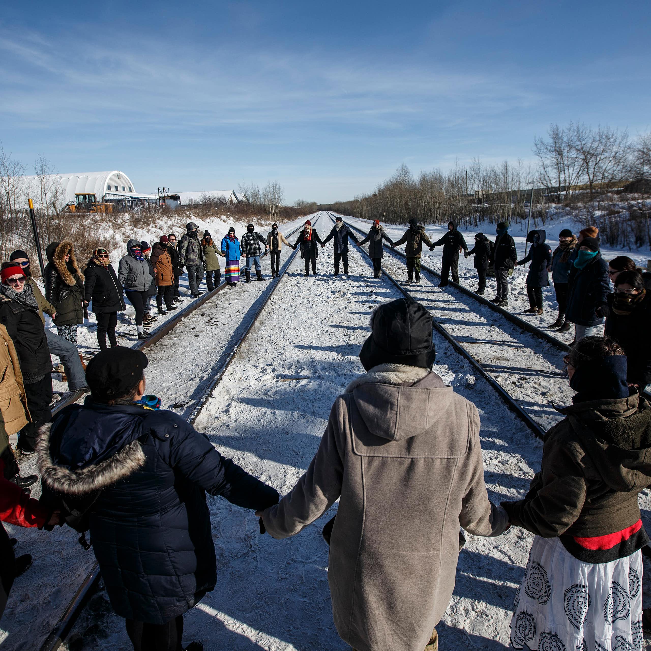 A group of people hold hands in a circle around a train track. 