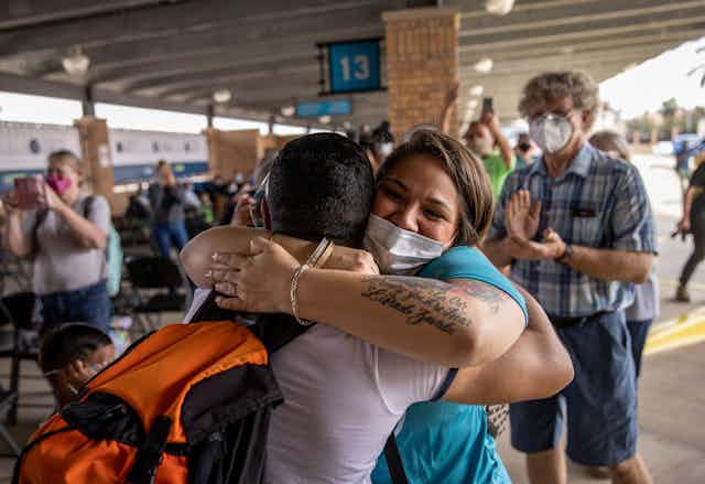 Woman in blue wearing a face mask hugs a person tightly while people clap in the background, at what appears to be a bus terminal