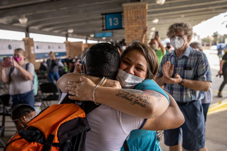 Woman in blue wearing a face mask hugs a person tightly while people clap in the background, at what appears to be a bus terminal