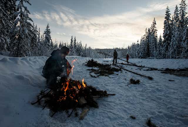 A person crouches near a fire in the snow near a road and surrounded by tall trees
