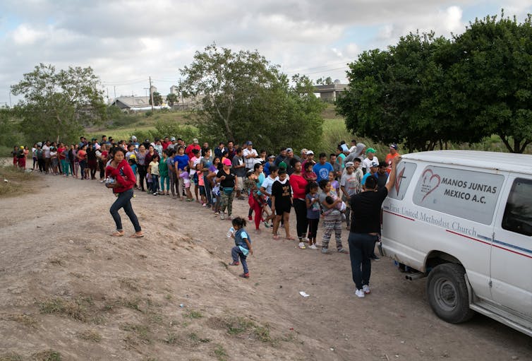 Line of people standing behind a van, with children playing in dirt in foreground