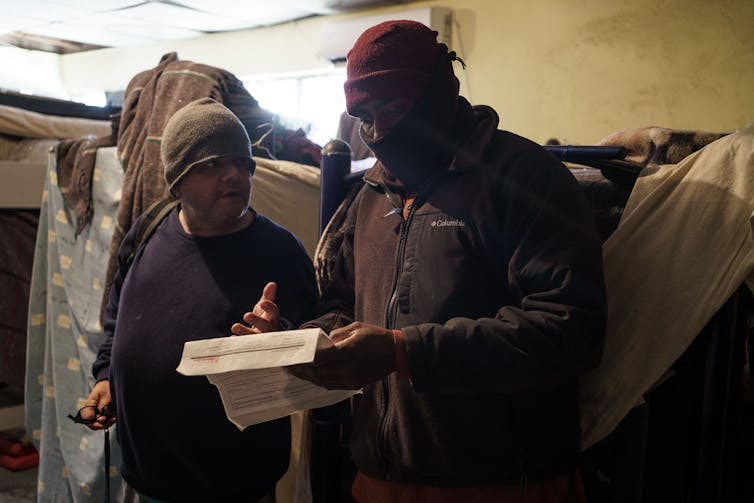 Two men in winter clothing in a room of bunk beds discuss a piece of paper that one man is holding