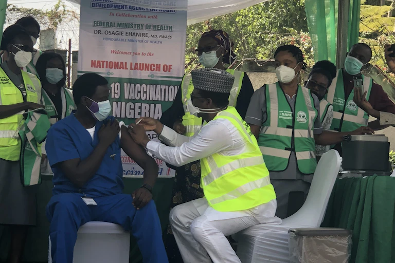 Man in hospital scrubs and face mask receives injection in his arm from another man in official waistcoat and mask, as others in similar uniforms look on