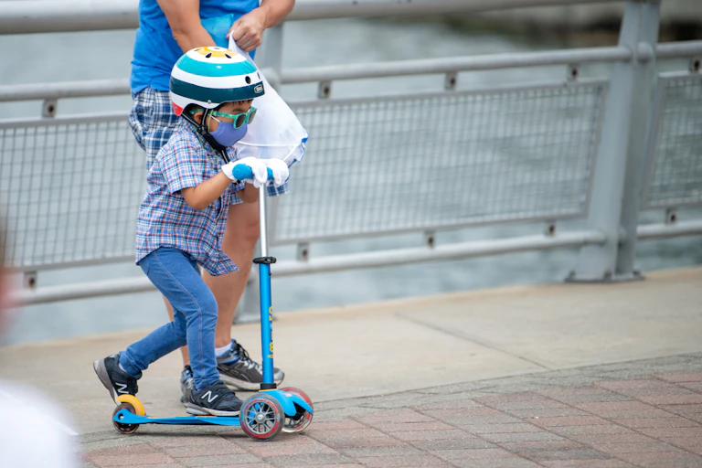 A little boy wearing a face mask, sunglasses and helmet rides a scooter next to his mom