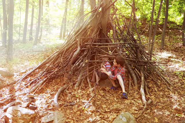 Two little boys in a forest sitting inside their den made of branches and twigs.