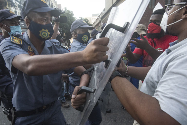 A row of police hold plastic shields up to barrier a row of protesters, all wearing face masks.