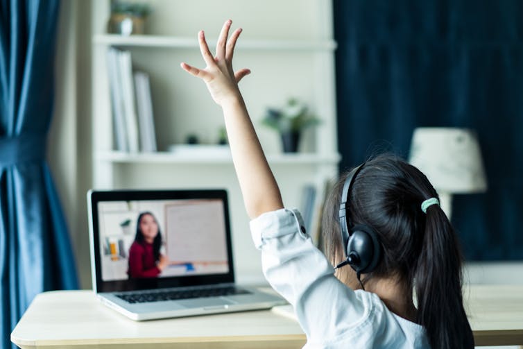 Child at laptop wearing headphones raises her hand