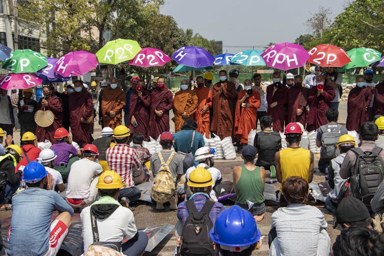 Men in hardhats sit on the ground facing monks in robes holding colorful umbrellas