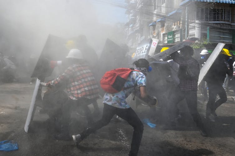 People with shields and hard hats duck and run for cover on a smoky street
