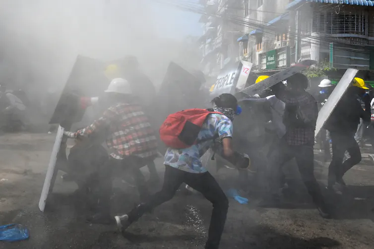 People with shields and hard hats duck and run for cover on a smoky street