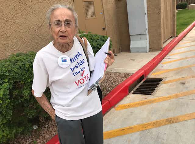 A woman with gray hair and glasses holds a clipboard and pen