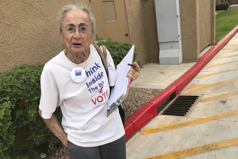 A woman with gray hair and glasses holds a clipboard and pen