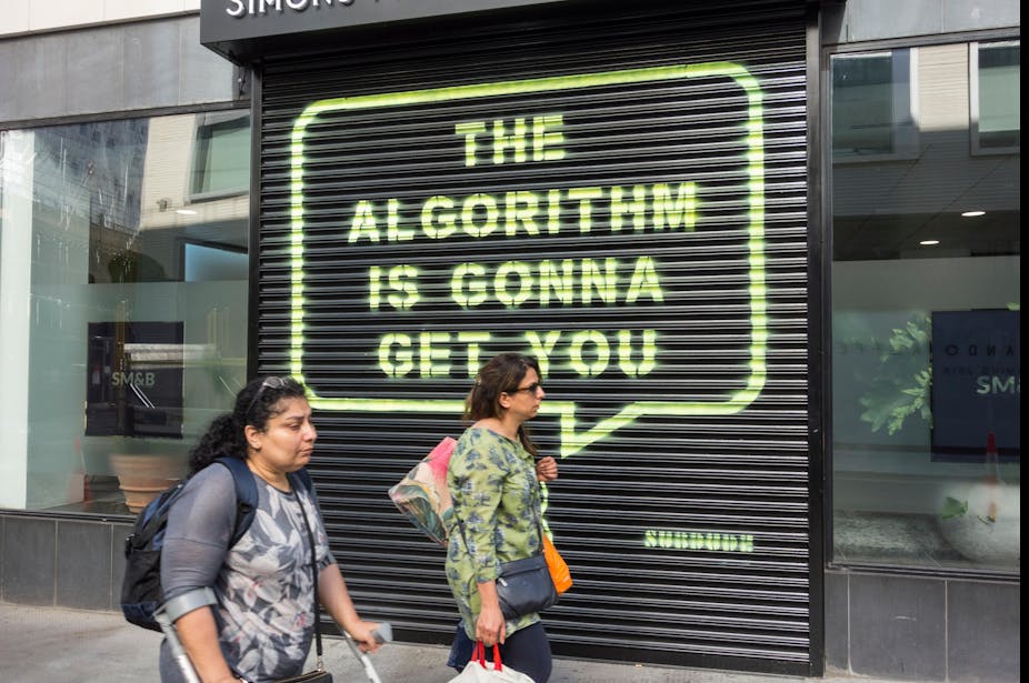 Woman in crutches and woman with bags walk past black shutter spraypainted in yellow with the words 'the algorithm is gonna get you' in a speech bubble