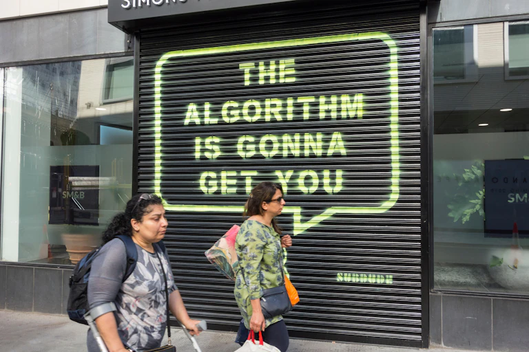 Woman in crutches and woman with bags walk past black shutter spraypainted in yellow with the words 'the algorithm is gonna get you' in a speech bubble
