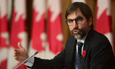 Heritage Minister Steven Guilbeault speaks with a row of Canadian flags behind him.