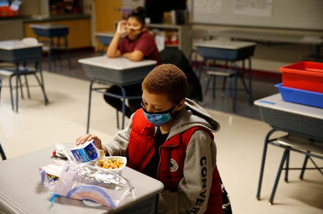 A school boy wearing a mask eats breakfast at his desk. 