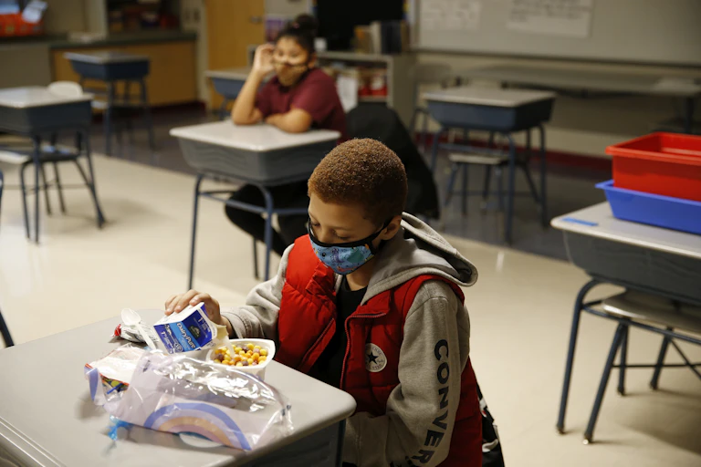 A school boy wearing a mask eats breakfast at his desk.