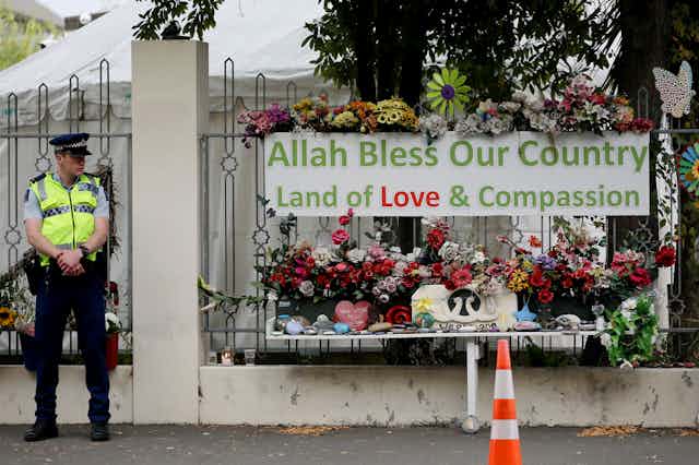 Police officer outside gate with sign reading 'Allah bless our country'