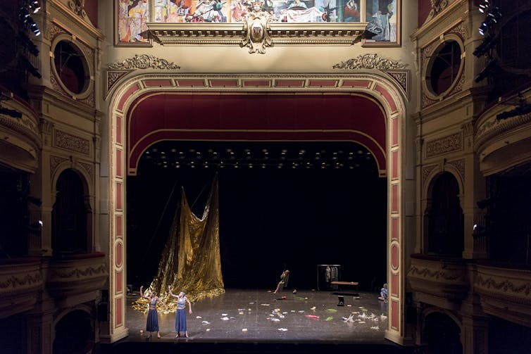 A wide shot of a proscenium arch. Two women look tiny against a messy stage.