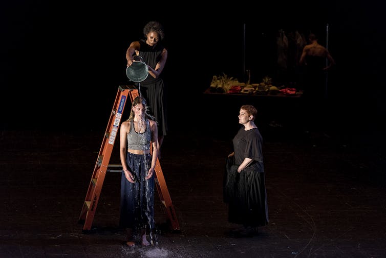 Three women, one pours a bucket of water over another.