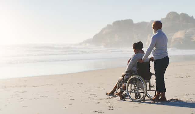 A woman sitting on a wheelchair with a man behind her holding the chair's handles. They're at a beach.