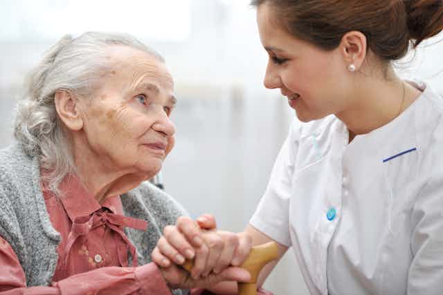 Elderly woman looking at young woman carer who is holding her hand.
