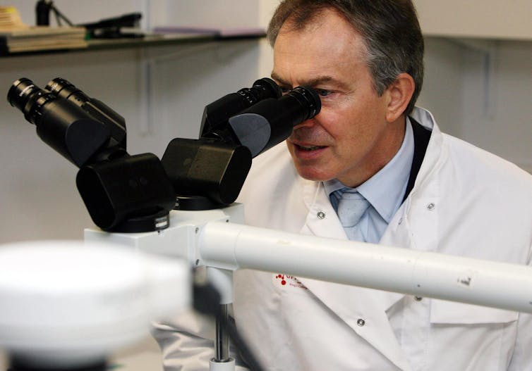 Tony Blair in a lab coat looking through a microscope.
