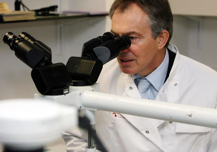 Tony Blair in a lab coat looking through a microscope.
