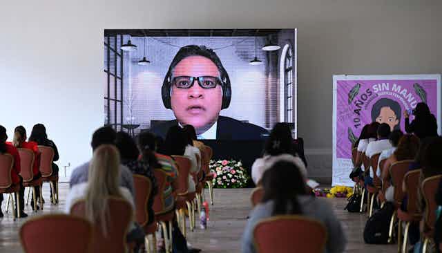 Backs of women sitting in chairs at a safe social distance watching a large screen. On the screen, a man speaks. Next to the screen is a banner reading '10 years without Manuela' 