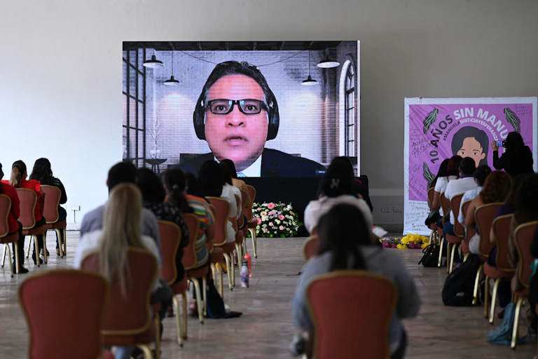 Backs of women sitting in chairs at a safe social distance watching a large screen. On the screen, a man speaks. Next to the screen is a banner reading '10 years without Manuela'