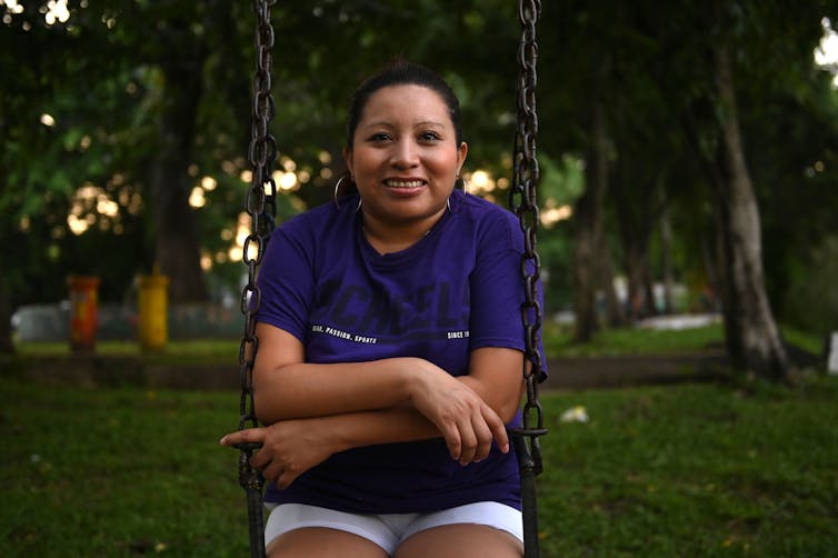 Woman in a purple T-shirt sits on a swing, smiling with arms crossed