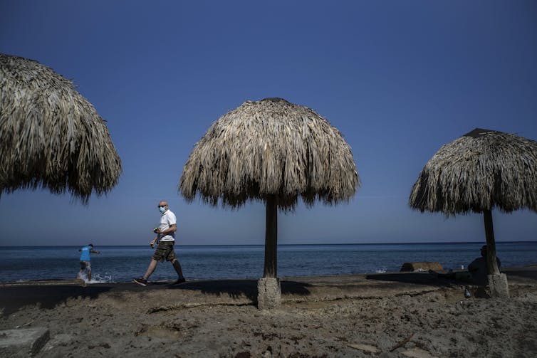 A man walks among beach huts wearing a mask.