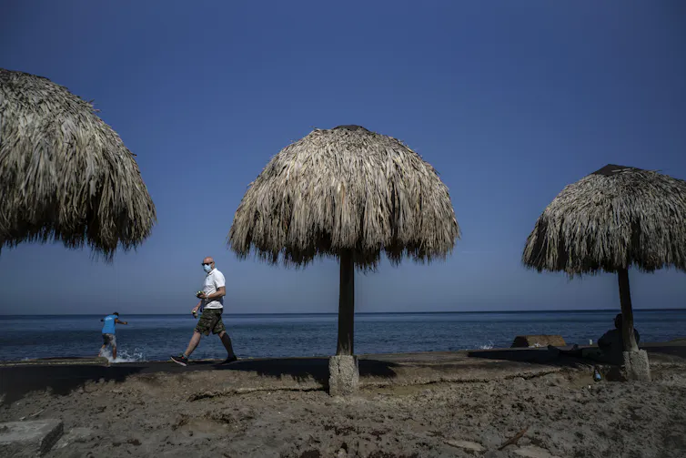 A man walks among beach huts wearing a mask.