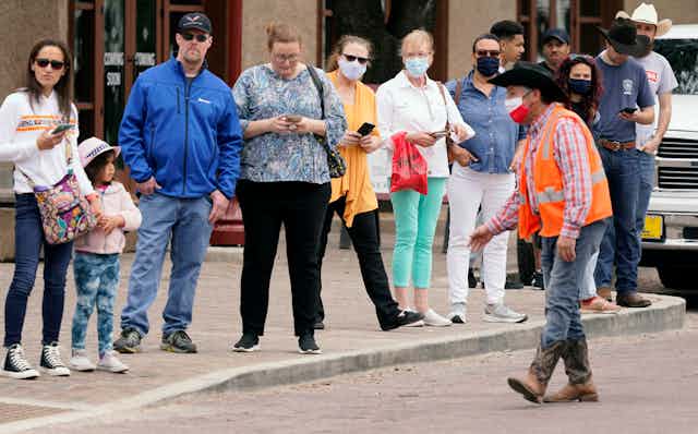 People stand on the curb very close to one another, some wearing masks, some not, as a man wearing a cowboy hat motions to them