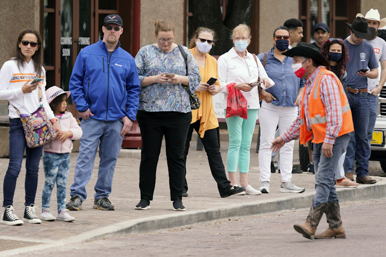 People stand on the curb very close to one another, some wearing masks, some not, as a man wearing a cowboy hat motions to them