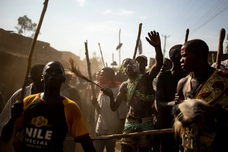 A throng of men in a village holding sticks with a young man in the middle of them, wearing animal skins, his face covered in clay.
