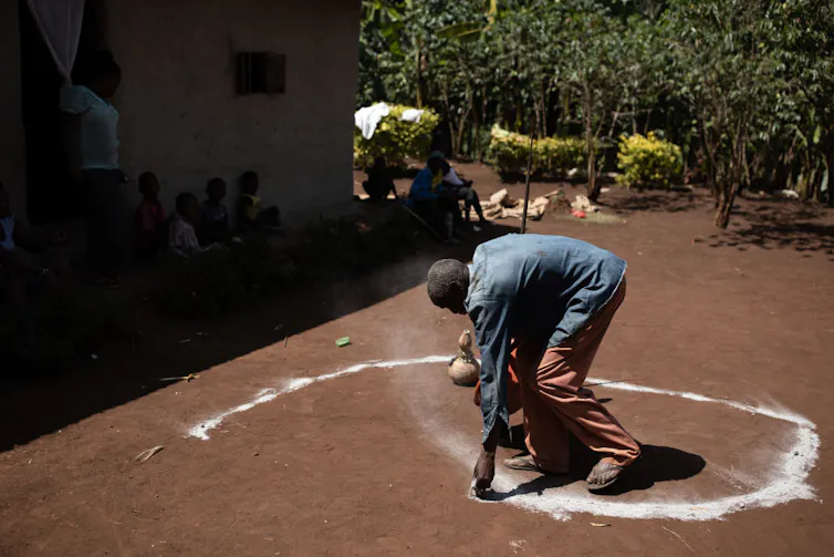 An elderly man is bent over, using white chalk to form a spiral shape on the dirt ground.
