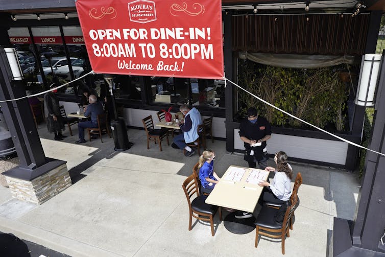 A sign over a restaurant in Los Angeles says 'Open for dine-in! 8:00AM to 8:00PM. Welcome Back!' above several diners at tables outside.