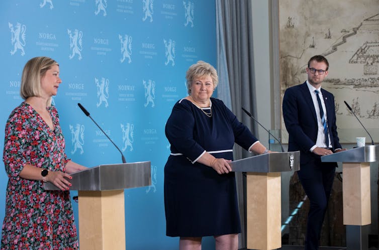 Two women and a man stand on a stage at lecterns, at a safe social distance.