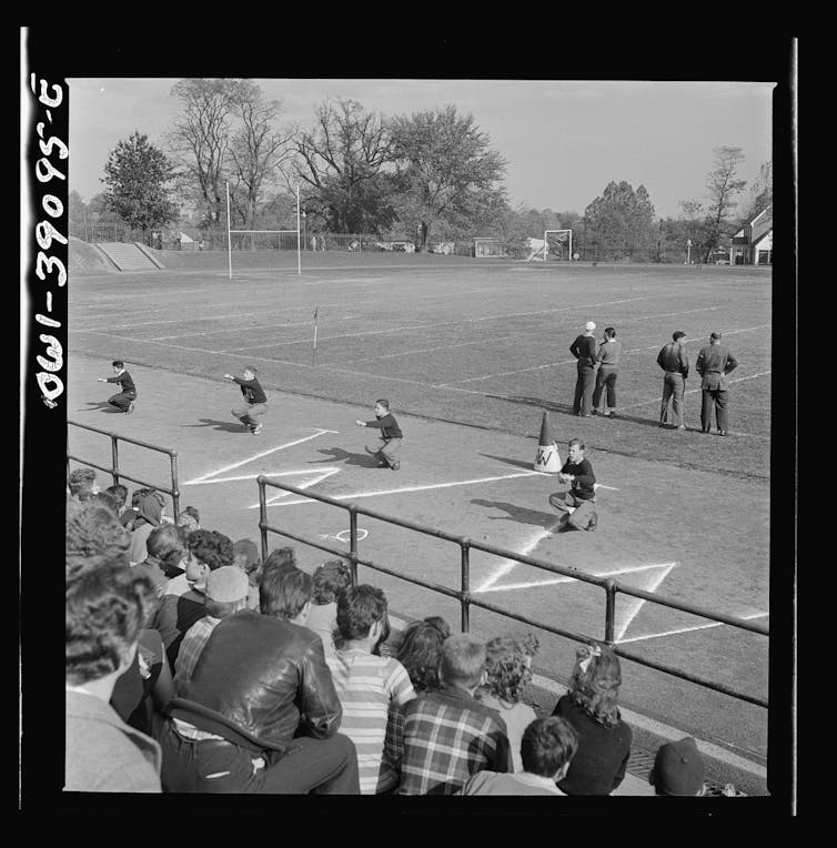Black and white photo. Four boys squat in a cheer in front of bleachers.