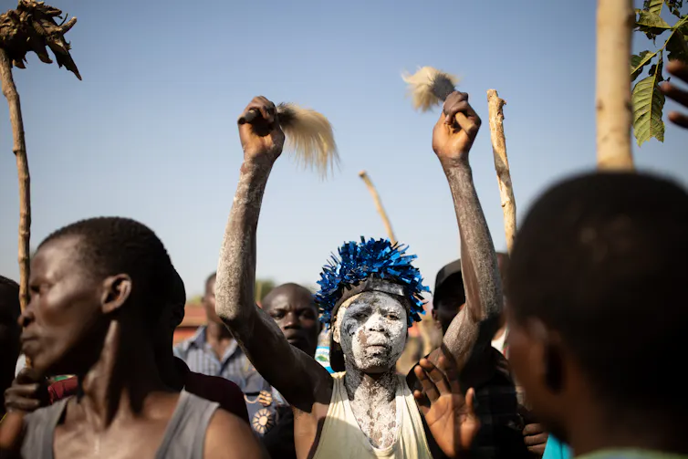 A crowd of men, in the centre a young man holding two cow's tail whisks in the air, white clay covering his face and a shiny blue piece of headgear.