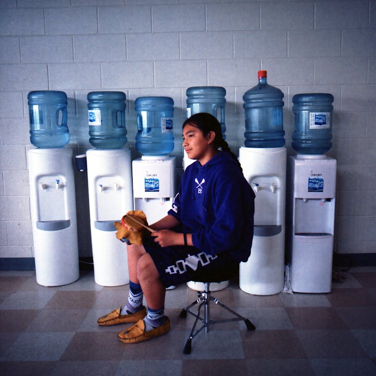 A young man with lacrosse stick in front of bottled water jugs.