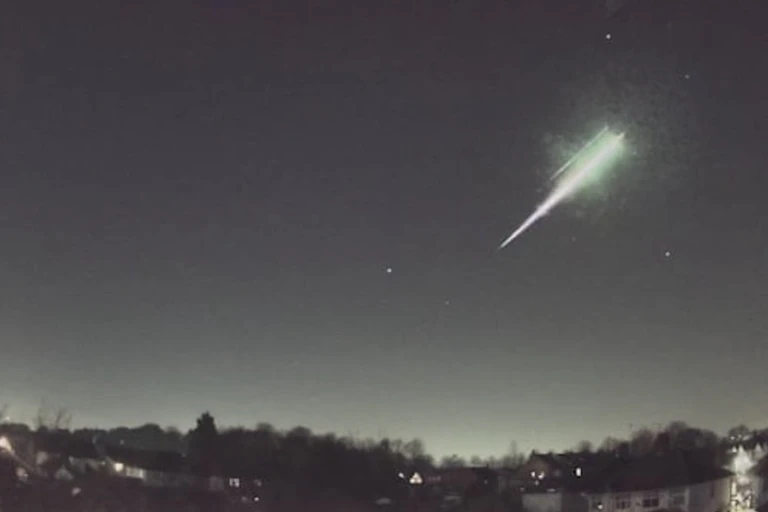 An image showing a bright streak moving through the night sky, with houses and trees below.