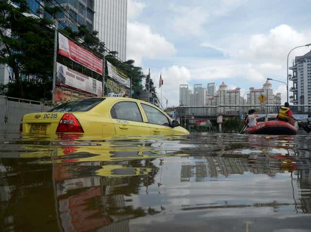 A yellow taxi on flooded street, large buildings in background