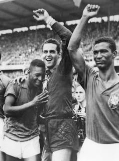 Um homem abraça dois jogadores de futebol, com os braços erguidos em vitória.