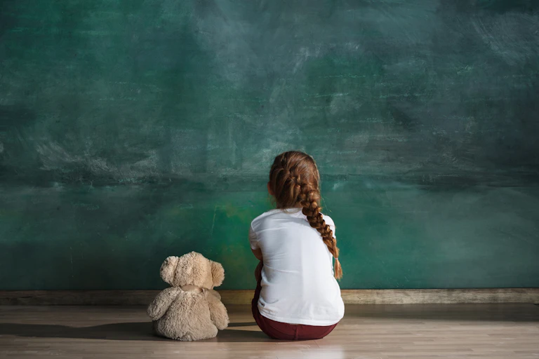 Young girl facing away from the camera sitting on the floor beside her teddy bear.