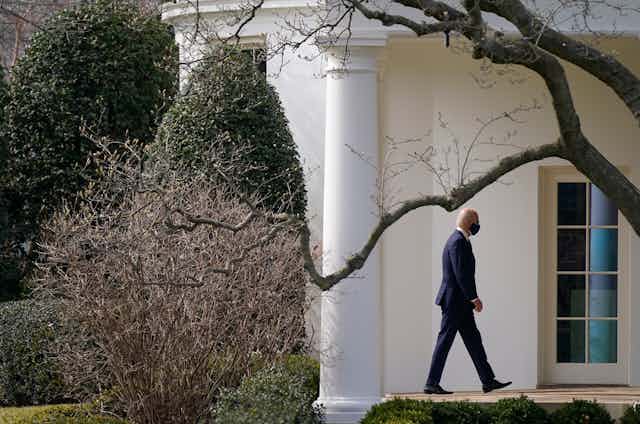Biden walks towards the Oval Office framed by trees and shrubs.