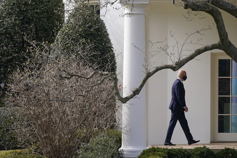 Biden walks towards the Oval Office framed by trees and shrubs.